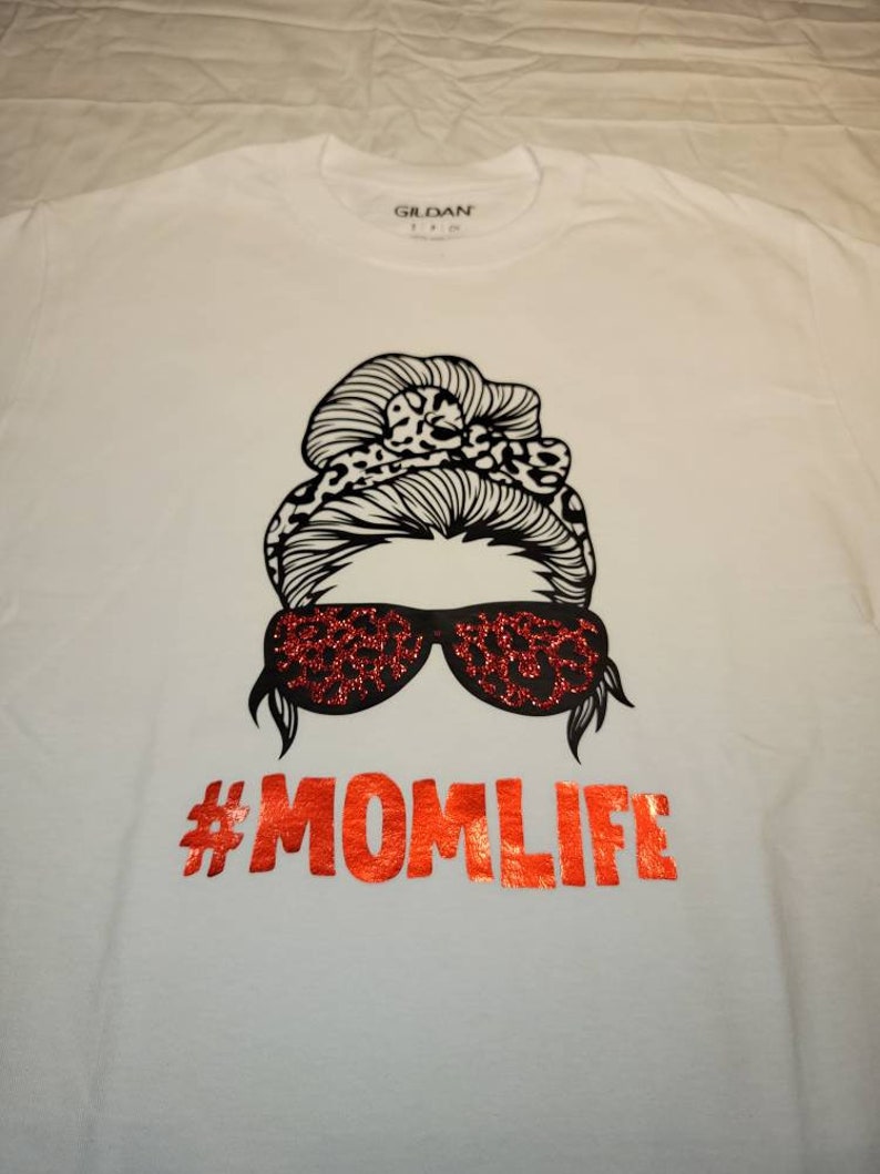 Peut inclure: T-shirt blanc avec un motif noir d'une femme avec un chignon d&eacute;sordonn&eacute;, des lunettes de soleil et le texte "#MOMLIFE" en paillettes rouges.