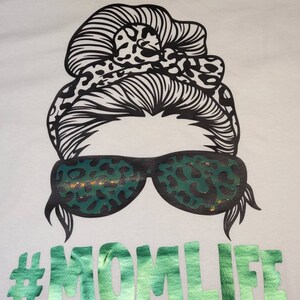 Peut inclure: T-shirt blanc avec un graphique noir et blanc d'une femme avec un chignon d&eacute;sordonn&eacute;, des lunettes de soleil et un imprim&eacute; l&eacute;opard. Le texte "#MOMLIFE" est imprim&eacute; en paillettes vertes.