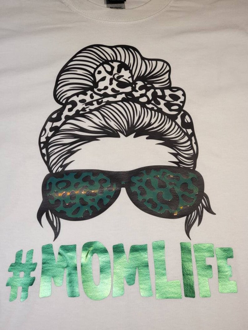 Peut inclure: T-shirt blanc avec un graphique noir et blanc d'une femme avec un chignon d&eacute;sordonn&eacute;, des lunettes de soleil et un imprim&eacute; l&eacute;opard. Le texte "#MOMLIFE" est imprim&eacute; en paillettes vertes.