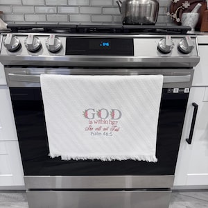 May include: A white kitchen towel with a fringed edge hangs on a stainless steel oven. The towel features the text "GOD is within her She will not fail Psalm 46:5" in a floral design. A stainless steel teapot sits on the stovetop.