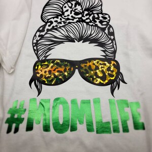 Peut inclure: T-shirt blanc avec un motif noir et blanc d'une femme avec un chignon d&eacute;sordonn&eacute;, un bandeau &agrave; imprim&eacute; l&eacute;opard et des lunettes de soleil. Les lunettes de soleil sont &eacute;galement &agrave; imprim&eacute; l&eacute;opard. Le texte "#MOMLIFE" est en vert sous le motif.