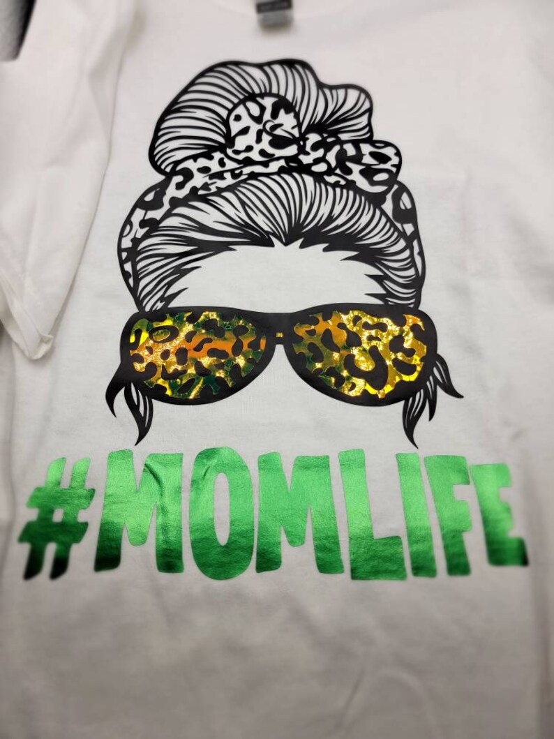 Peut inclure: T-shirt blanc avec un motif noir et blanc d'une femme avec un chignon d&eacute;sordonn&eacute;, un bandeau &agrave; imprim&eacute; l&eacute;opard et des lunettes de soleil. Les lunettes de soleil sont &eacute;galement &agrave; imprim&eacute; l&eacute;opard. Le texte "#MOMLIFE" est en vert sous le motif.