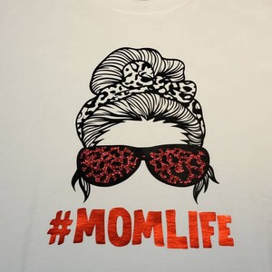 Peut inclure: T-shirt blanc avec une illustration en noir et blanc d'une femme avec un chignon d&eacute;sordonn&eacute; et des lunettes de soleil. Les lunettes de soleil et le chignon ont un motif l&eacute;opard. Le texte "#MOMLIFE" est en paillettes rouges.