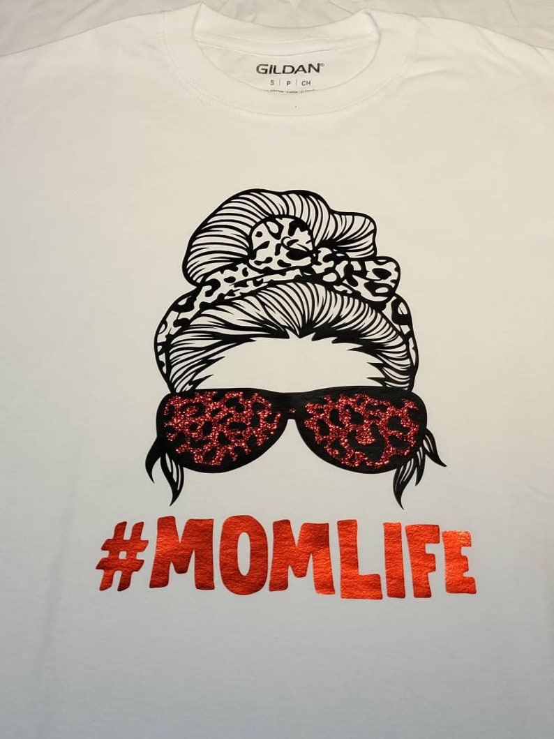 Peut inclure: T-shirt blanc avec une illustration en noir et blanc d'une femme avec un chignon d&eacute;sordonn&eacute; et des lunettes de soleil. Les lunettes de soleil et le chignon ont un motif l&eacute;opard. Le texte "#MOMLIFE" est en paillettes rouges.