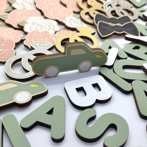 May include: A collection of wooden craft supplies, including letters, a truck, rings, flowers, and a bow tie. The letters are in green and white, and the truck is green with white wheels. The rings are silver with a diamond accent. The flowers are pink and white. The bow tie is black.