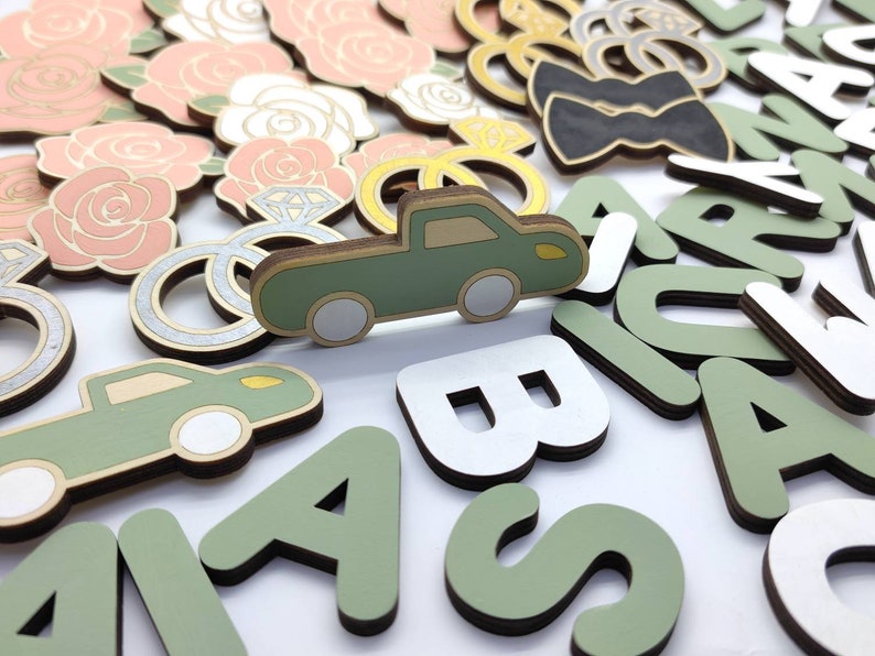 May include: A collection of wooden craft supplies, including letters, a truck, rings, flowers, and a bow tie. The letters are in green and white, and the truck is green with white wheels. The rings are silver with a diamond accent. The flowers are pink and white. The bow tie is black.