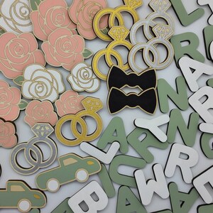 May include: A collection of wooden craft supplies featuring pink and white roses, black bow ties, gold and silver rings, and green and white letters.