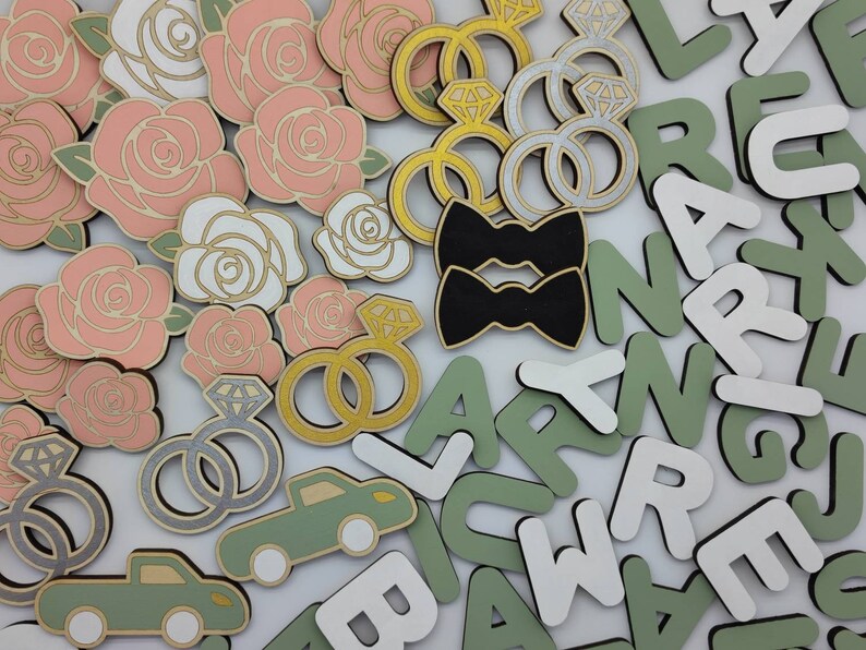 May include: A collection of wooden craft supplies featuring pink and white roses, black bow ties, gold and silver rings, and green and white letters.