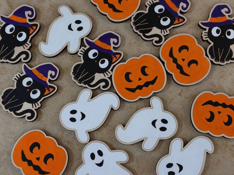 May include: A collection of Halloween-themed wooden ornaments featuring pumpkins, ghosts, and black cats. The ornaments are painted in orange, white, and black.