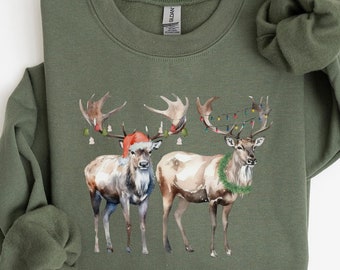 May include: A green sweatshirt with a Christmas-themed design featuring two deer with antlers, one wearing a Santa hat and the other with a wreath around its neck. The deer are surrounded by Christmas lights and greenery.