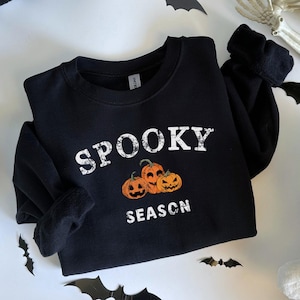 May include: Black sweatshirt with the words "Spooky Season" and a graphic of three jack-o'-lanterns. The sweatshirt is surrounded by Halloween decorations, including bats, a skeleton, and ghost-shaped pillows. A black and white checkered pillow is also in the image.