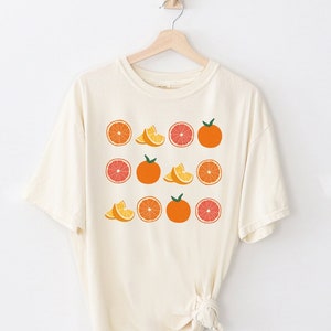 以下が含まれることがあります： オレンジとオレンジのスライスがグラフィックプリントされた白いTシャツ。グラフィックはオレンジ、ピンク、イエローのカラーで構成されています。