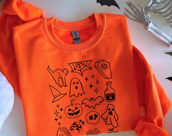 Könnte beinhalten: Ein orangefarbenes Halloween-Sweatshirt mit schwarzen grafischen Illustrationen einer Katze, eines Spinnennetzes, eines Geistes, eines Kürbisses und anderer gruseliger Symbole. Das Sweatshirt ist von Halloween-Dekorationen umgeben.