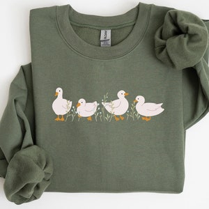Puede incluir: Una sudadera verde con un gráfico de cinco patos blancos en fila. Los patos están mirando hacia el lado derecho de la imagen. La sudadera tiene cuello redondo y mangas largas. El texto "EMMA & ANNA" está impreso en negro en la parte inferior de la imagen.