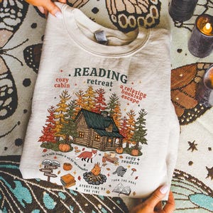 Op de afbeelding: Een cr&egrave;mekleurige sweatshirt met een afbeelding van een hut in een bos, pompoenen en de tekst "Reading Retreat". Het ontwerp bevat een vos, een kampvuur en de teksten "cozy cabin" en "a relaxing bookish escape".