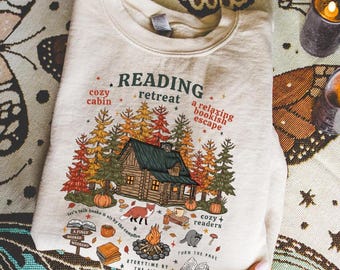May include: A cream-coloured sweatshirt featuring a cabin graphic in a forest setting, with pumpkins and the words "Reading Retreat." The design includes a fox, campfire, and the text "cozy cabin" and "a relaxing bookish escape."