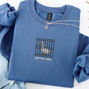 Puede incluir: Una sudadera azul con un gráfico de un ganso blanco en una jaula y el texto "got too silly".