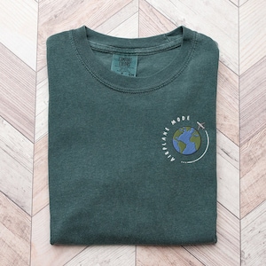Op de afbeelding: Een donkergroen T-shirt met een wit geborduurd ontwerp van een wereldbol met een vliegtuig dat eromheen vliegt. De tekst "Airplane Mode" staat rond de wereldbol.