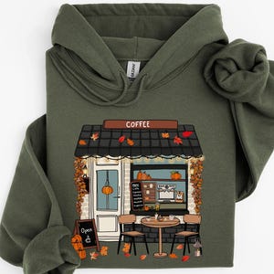 May include: An olive green hoodie with a coffee shop graphic. The shop has a black roof, a door with a pumpkin, and a sign that says "Open". The word "Coffee" is above the shop. Autumn leaves and pumpkins decorate the scene.
