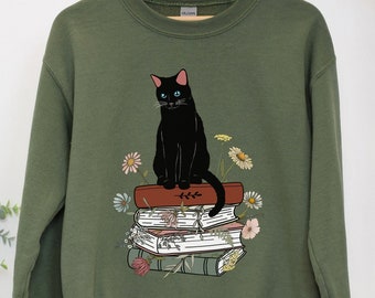 May include: A green sweatshirt with a black cat sitting on a stack of books. The cat has blue eyes and is surrounded by flowers.