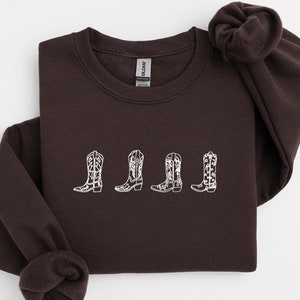 Puede incluir: Una sudadera marrón con un diseño bordado blanco de cuatro botas de vaquero.