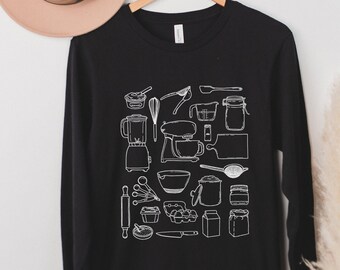 May include: Black long-sleeve shirt with a white outline of various kitchen tools and appliances, including a blender, mixer, rolling pin, measuring cups, and more.