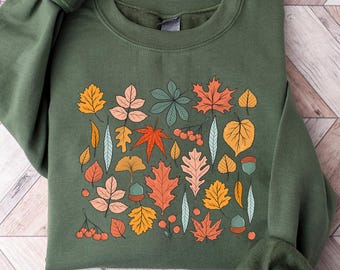 Puede incluir: Sudadera verde con un diseño de hojas de otoño coloridas. Las hojas son en tonos naranja, amarillo, rosa y verde. La sudadera tiene mangas largas y cuello redondo. El texto "EMMA & ANNA" está en la parte inferior.