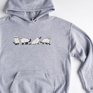 Puede incluir: Una sudadera con capucha gris con un gr&aacute;fico de cuatro gatos blancos en la parte delantera. Los gatos est&aacute;n en l&iacute;nea, y el del medio est&aacute; sentado. El texto "EMMA & ANNA" est&aacute; impreso en la esquina inferior derecha de la sudadera.