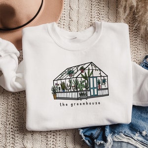 May include: White sweatshirt with a greenhouse illustration in black and green, and the text "the greenhouse". The image shows the sweatshirt with denim shorts and a beige hat. The design features a variety of plants.