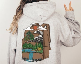 Può includere: Una felpa grigia con una grafica di uno zaino marrone con una scena di montagna al suo interno. La scena include un lago, alberi, una tenda e una cascata. Il testo "EMMA & ANNA" è stampato nell'angolo in basso a destra della felpa.