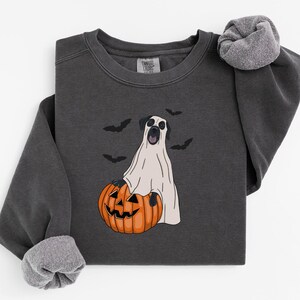 Può includere: Una felpa girocollo grigio antracite con la grafica di un cane travestito da fantasma, seduto dietro una zucca arancione. Il disegno include piccole sagome di pipistrelli neri che circondano il cane sul tessuto scuro.