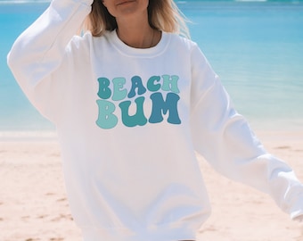 Può includere: Felpa bianca con una grafica blu e verde che dice "Beach Bum".