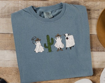 May include: A light blue t-shirt with a graphic of four ghosts in a line. The ghosts are wearing cowboy hats and one is holding a lasso. There is a cactus between two of the ghosts. The text "Emma & Anna" is printed on the bottom right corner of the image.