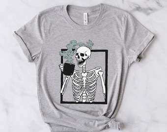 May include: A grey t-shirt featuring a black and white illustration of a skeleton holding a steaming cup of coffee. The skeleton is framed in a black rectangle.