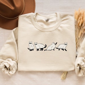 Puede incluir: Una sudadera de color crema con un gráfico de cinco gatos vestidos de fantasmas. Los gatos están en fila, caminando de izquierda a derecha. La sudadera tiene mangas largas y cuello redondo.