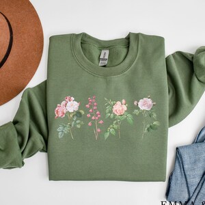 May include: A green sweatshirt with a floral print of pink and white roses and green leaves. The sweatshirt has a crew neck and long sleeves.