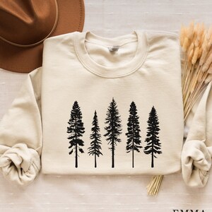 May include: A cream-colored sweatshirt with a black silhouette of five pine trees printed on the front. The sweatshirt has long sleeves and a crew neck.