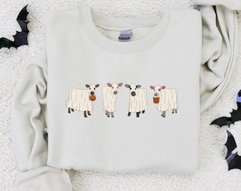 May include: A light beige sweatshirt with a graphic of four cows dressed as ghosts holding buckets of candy. The cows are white with black spots and have a black nose and eyes. The sweatshirt has a crew neck and long sleeves.