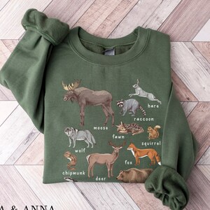 May include: A green sweatshirt with a graphic print of various forest animals, including a moose, wolf, deer, fox, squirrel, chipmunk, raccoon, hare, and fawn. The animals are labeled with their names.