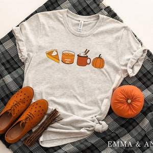 May include: A gray t-shirt with a pumpkin spice latte, pumpkin pie, and a pumpkin graphic. The shirt is laying on a black and white plaid blanket with two brown shoes and a brown pumpkin.