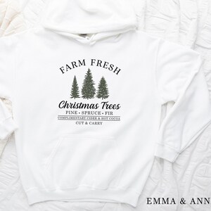 Op de afbeelding: Witte hoodie met een zwarte afbeelding met de tekst "Farm Fresh" met een afbeelding van drie kerstbomen en de tekst "Christmas Trees Pine Spruce Fir Complimentary Cider & Hot Cocoa Cut & Carry"