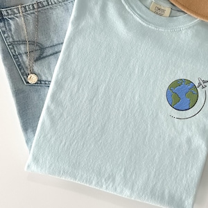 May include: A light blue t-shirt with an embroidered design of a globe with a plane flying around it. The text "Comfort Colors" is visible on the tag.
