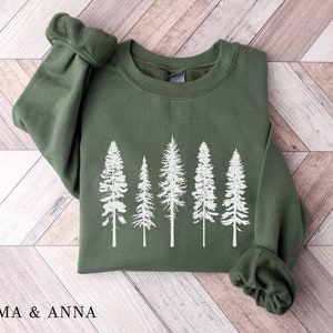 Puede incluir: Una sudadera verde con una silueta blanca de una fila de pinos impresa en la parte delantera. La sudadera est&aacute; doblada y colocada sobre una superficie de madera.