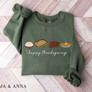 May include: Green sweatshirt with the words "happy thanksgiving" in white script. The sweatshirt features illustrations of Thanksgiving food: mashed potatoes, casserole, a turkey, and a pie. The brand name "EMMA & ANNA" is at the bottom.
