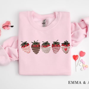 Puede incluir: Sudadera rosa con un gr&aacute;fico de cinco fresas ba&ntilde;adas en chocolate y chocolate blanco. Las fresas tienen corazones rojos en el centro. La sudadera tiene cuello redondo y mangas largas.