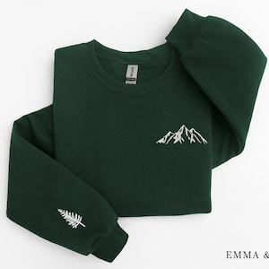 Könnte beinhalten: Ein dunkelgrüner Pullover mit einem weißen gestickten Bergmotiv auf der Vorderseite und einem weißen gestickten Kiefernbaum auf dem linken Ärmel. Der Pullover hat einen Rundhalsausschnitt und lange Ärmel.