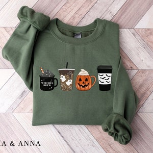 Puede incluir: Una sudadera verde con un diseño de temática de Halloween que presenta cuatro tazas de café. Las tazas tienen diferentes diseños, incluyendo una calabaza, un fantasma y una taza que dice "Witches Brew".