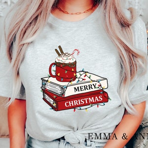 May include: A gray t-shirt with a red mug of hot chocolate topped with whipped cream and a candy cane. The mug is surrounded by colorful Christmas lights and a stack of books with the text "Merry Christmas".