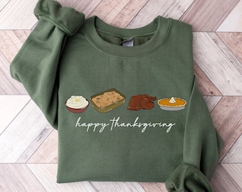 May include: Green sweatshirt with the words "happy thanksgiving" in white script. The sweatshirt features illustrations of Thanksgiving food: mashed potatoes, casserole, a turkey, and a pie. The brand name "EMMA & ANNA" is at the bottom.