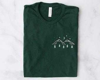 Puede incluir: Una camiseta verde oscuro con un diseño bordado en blanco de dos montañas, una luna creciente y estrellas. El diseño está centrado en la parte delantera de la camiseta.